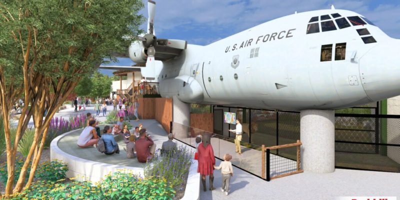 Architectural rendering of a US Air Force C-130 aircraft mounted above a zoo lion habitat, with visitors seated at an amphitheater below.
