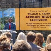 A speaker at a podium addresses a seated crowd in front of a large banner reading Future home of William S. Smith African Wilds: Carnivores.