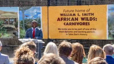 A speaker at a podium addresses a seated crowd in front of a large banner reading Future home of William S. Smith African Wilds: Carnivores.