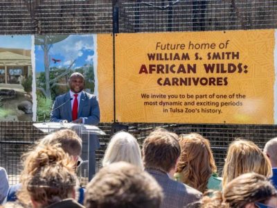 A speaker at a podium addresses a seated crowd in front of a large banner reading Future home of William S. Smith African Wilds: Carnivores.