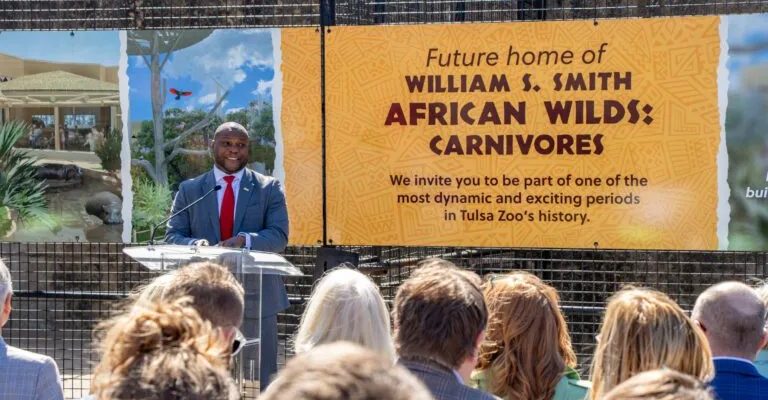 A speaker at a podium addresses a seated crowd in front of a large banner reading Future home of William S. Smith African Wilds: Carnivores.