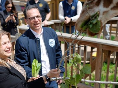 Governor Josh Shapiro and First Lady Lori Shapiro hold leafy browse up to a giraffe leaning over a wooden railing.