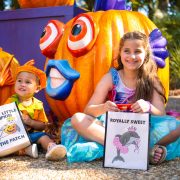 Two costumed children sit in front of large jack-o'-lanterns carved and painted as tropical fish, holding trick-or-treat bags.