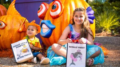 Two costumed children sit in front of large jack-o'-lanterns carved and painted as tropical fish, holding trick-or-treat bags.