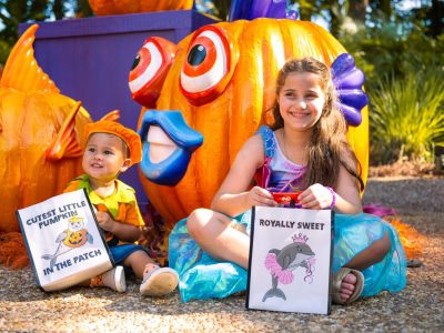 Two costumed children sit in front of large jack-o'-lanterns carved and painted as tropical fish, holding trick-or-treat bags.