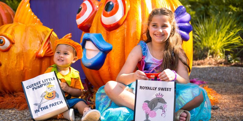 Two costumed children sit in front of large jack-o'-lanterns carved and painted as tropical fish, holding trick-or-treat bags.