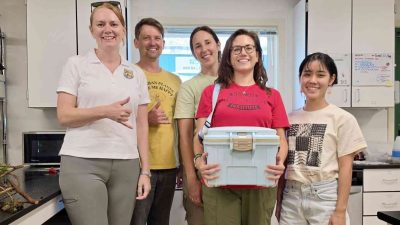 Five staff members stand together in a laboratory, one holding a white transport cooler containing rescued snails.