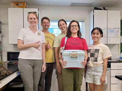 Five staff members stand together in a laboratory, one holding a white transport cooler containing rescued snails.