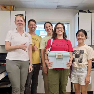 Five staff members stand together in a laboratory, one holding a white transport cooler containing rescued snails.