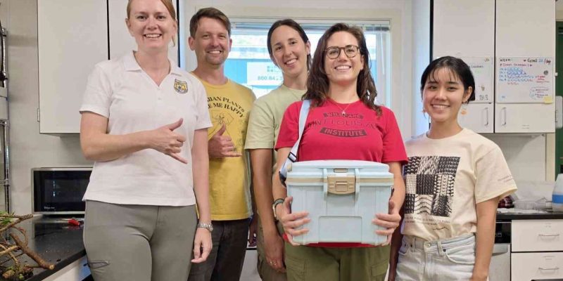 Five staff members stand together in a laboratory, one holding a white transport cooler containing rescued snails.