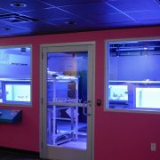 A darkened exhibit room with magenta walls and two large windows looking into a blue-lit laboratory holding coral tanks.