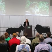 A woman speaks at a podium in front of a seated audience, with lion silhouettes and a projected lion image behind her.