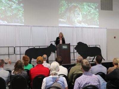 A woman speaks at a podium in front of a seated audience, with lion silhouettes and a projected lion image behind her.