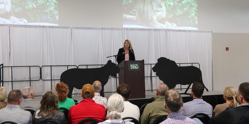 A woman speaks at a podium in front of a seated audience, with lion silhouettes and a projected lion image behind her.