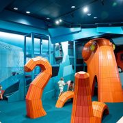 Children climb through a large orange octopus-shaped play structure inside a blue-walled indoor discovery center.