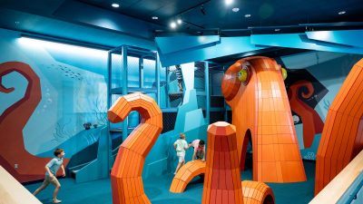 Children climb through a large orange octopus-shaped play structure inside a blue-walled indoor discovery center.