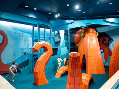 Children climb through a large orange octopus-shaped play structure inside a blue-walled indoor discovery center.