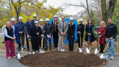 A row of people in white hard hats hold gold shovels over a mound of soil at the Wild Iowa groundbreaking, autumn trees behind them.