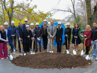 A row of people in white hard hats hold gold shovels over a mound of soil at the Wild Iowa groundbreaking, autumn trees behind them.