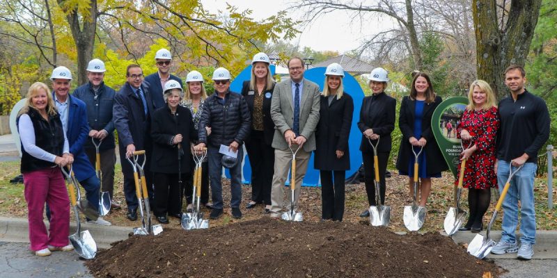 A row of people in white hard hats hold gold shovels over a mound of soil at the Wild Iowa groundbreaking, autumn trees behind them.