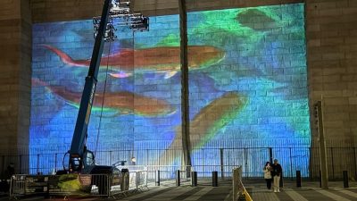 A blue and green undersea scene projected across the stone masonry of the Brooklyn Bridge at night, with orange fish shapes across the center, a telescopic lift carrying the projector rig at the left, and two people walking past barriers at the base.