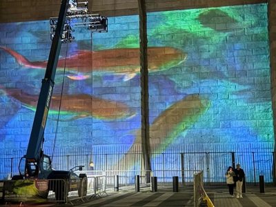 A blue and green undersea scene projected across the stone masonry of the Brooklyn Bridge at night, with orange fish shapes across the center, a telescopic lift carrying the projector rig at the left, and two people walking past barriers at the base.