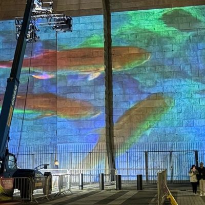 A blue and green undersea scene projected across the stone masonry of the Brooklyn Bridge at night, with orange fish shapes across the center, a telescopic lift carrying the projector rig at the left, and two people walking past barriers at the base.