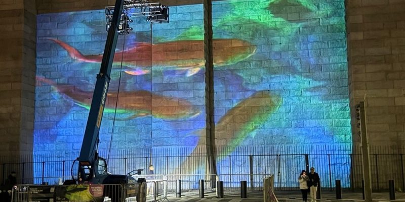 A blue and green undersea scene projected across the stone masonry of the Brooklyn Bridge at night, with orange fish shapes across the center, a telescopic lift carrying the projector rig at the left, and two people walking past barriers at the base.