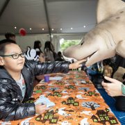 A boy in a black puffer jacket and glasses reaches out to touch the snout of a large shark puppet held across a table covered in an orange Halloween tablecloth.
