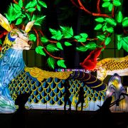 Visitors in silhouette walk past enormous illuminated lantern sculptures of deer-like creatures among glowing green foliage at night.