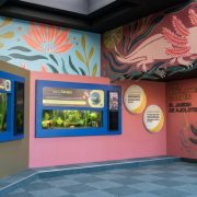 Digital rendering of a gallery painted coral pink and blue with large axolotl murals, planted aquarium tanks along one wall and two guests reading a sign.