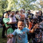 A child with a painted face reaches for floating bubbles in a crowd of families at an outdoor autumn event.