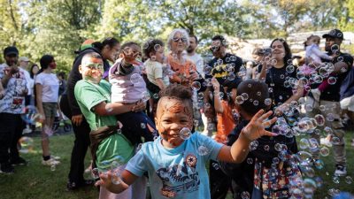 A child with a painted face reaches for floating bubbles in a crowd of families at an outdoor autumn event.