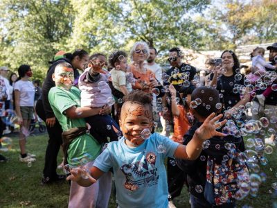A child with a painted face reaches for floating bubbles in a crowd of families at an outdoor autumn event.