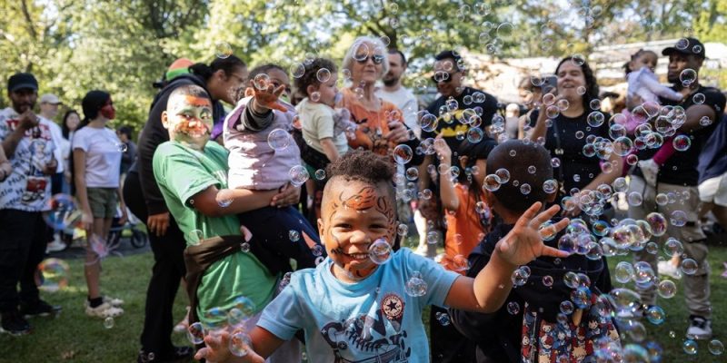 A child with a painted face reaches for floating bubbles in a crowd of families at an outdoor autumn event.