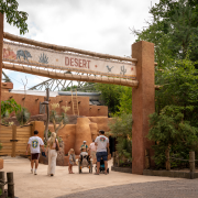 Visitors walk beneath a tall adobe-style gateway with a sign reading Desert, with cacti and planting on either side.