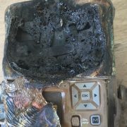 A camera trap housing destroyed by fire, its upper half melted into a mass of blackened bubbled plastic, with the scorched keypad and OK button still intact below.