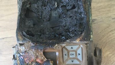 A camera trap housing destroyed by fire, its upper half melted into a mass of blackened bubbled plastic, with the scorched keypad and OK button still intact below.