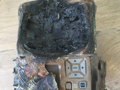 A camera trap housing destroyed by fire, its upper half melted into a mass of blackened bubbled plastic, with the scorched keypad and OK button still intact below.