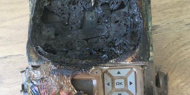 A camera trap housing destroyed by fire, its upper half melted into a mass of blackened bubbled plastic, with the scorched keypad and OK button still intact below.