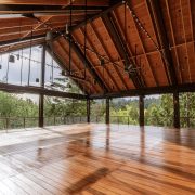 An open-sided pavilion with a high timber roof, hanging lights and a polished wood deck, looking out through steel posts to forested hills.