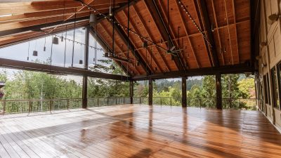 An open-sided pavilion with a high timber roof, hanging lights and a polished wood deck, looking out through steel posts to forested hills.