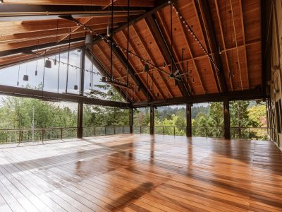 An open-sided pavilion with a high timber roof, hanging lights and a polished wood deck, looking out through steel posts to forested hills.