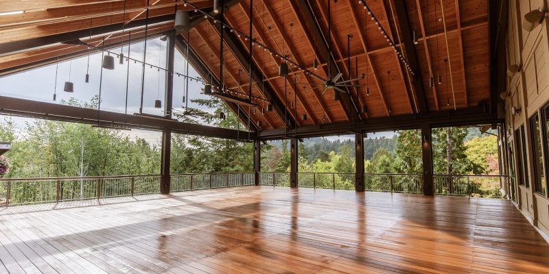 An open-sided pavilion with a high timber roof, hanging lights and a polished wood deck, looking out through steel posts to forested hills.