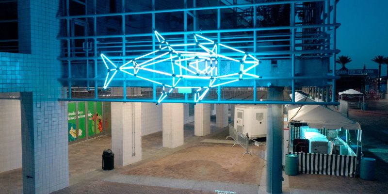 A large illuminated sculpture shaped like a tuna glows against a dark city street outside the entrance to Audubon Aquarium, its surface lit in shifting colors.