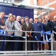 A group of officials stands on a metal platform beside a large blue combined heat and power unit, holding lengths of cut blue ribbon.