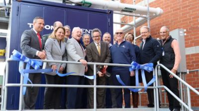 A group of officials stands on a metal platform beside a large blue combined heat and power unit, holding lengths of cut blue ribbon.