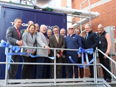 A group of officials stands on a metal platform beside a large blue combined heat and power unit, holding lengths of cut blue ribbon.
