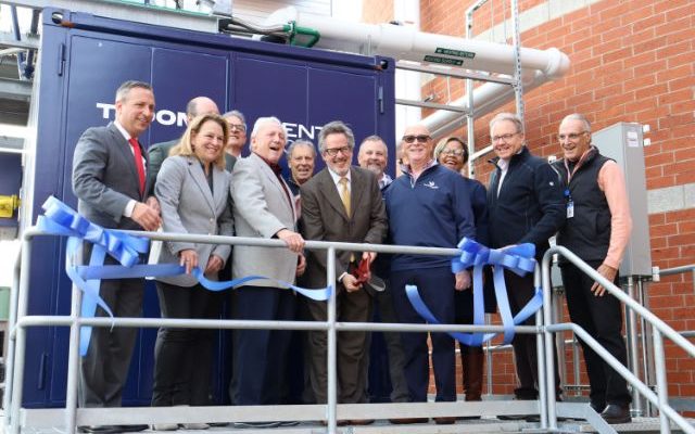 A group of officials stands on a metal platform beside a large blue combined heat and power unit, holding lengths of cut blue ribbon.