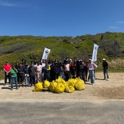 A large group of volunteers, most of them schoolchildren in warm clothing, stand on a gravel verge behind a pile of full yellow waste bags, with two banners and green coastal fynbos on the slope behind them.